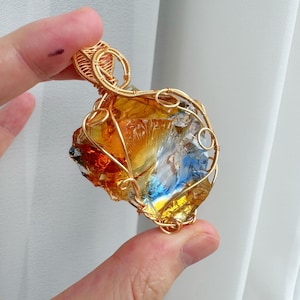 May include: A pendant featuring a raw, translucent amber and blue glass stone encased in intricate gold-coloured wire wrapping. The design includes a decorative wire bail at the top, securing the irregular, multi-coloured stone within a metal frame.