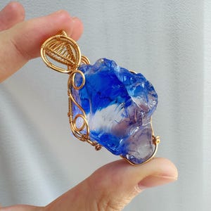 May include: A blue and clear crystal pendant with a gold wire wrap. The crystal has a rough, natural texture and is suspended from a gold bail. The pendant is held in a hand against a soft, neutral background.