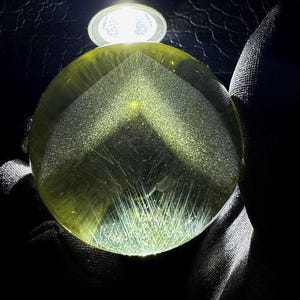 May include: A round, transparent, yellow-green gemstone with internal inclusions and a faceted top. The gem is illuminated by a bright light source, creating a glowing effect. The background is dark, emphasizing the gem's clarity and color.