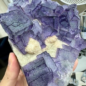 Puede incluir: Primer plano de una formación de cristales de fluorita, que muestra un tono púrpura intenso con secciones más claras y translúcidas. Los cristales tienen una estructura geométrica en capas. La base es de color beige claro y el aspecto general es natural.