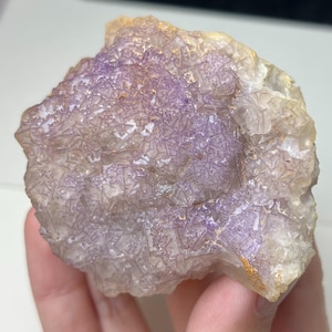 May include: A raw, natural fluorite specimen with a mix of purple and clear crystalline structures. The stone has a rough, textured surface with hints of yellow and gold.