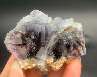 Könnte beinhalten: Natürliches Fluorid-Mineralpräparat mit einer Mischung aus violetten, weißen und klaren Kristallstrukturen. Die Basis hat eine beige und bräunliche Farbe. Das Präparat wird in einer Hand vor einem dunklen Hintergrund gehalten.