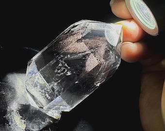 May include: A clear, faceted quartz crystal point featuring internal inclusions of dark, metallic-looking mineral deposits. The crystal has a hexagonal shape with sharp, defined edges and a polished, transparent surface that reflects light.