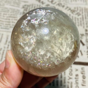 Peut inclure: Une boule de cristal sphérique transparente avec des inclusions irisées internes. La sphère a une surface lisse et réfléchit la lumière, affichant une gamme de couleurs. Le cristal est tenu dans une main.