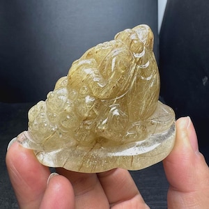 May include: A carved golden rutilated quartz figurine shaped like a three-legged toad. The translucent stone features visible needle-like golden inclusions throughout its polished surface, depicting the toad sitting on a flat, rounded base.