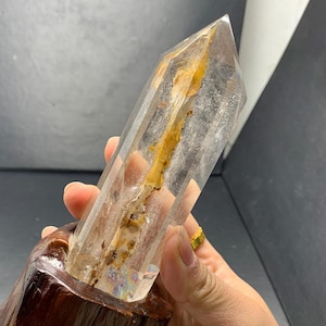 May include: A clear quartz crystal point with a brown inclusion inside. The crystal is sitting in a wooden base.
