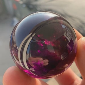 Op de afbeelding: Een gladde, bolvormige, doorschijnende paarse glazen bol die de omgeving reflecteert op het gepolijste oppervlak. De dieppaarse kleur gaat over in lichtere magentatinten waar het licht door het heldere materiaal heen schijnt.