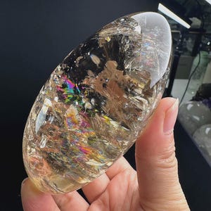 以下が含まれることがあります： 内部に亀裂やインクルージョンを含む、磨かれた楕円形のクリアクォーツクリスタル。石の透明な本体全体に鮮やかな虹色の輝きが見られ、内部には針状の暗い鉱物の堆積物が確認できます。
