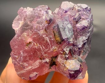Könnte beinhalten: Nahaufnahme eines Fluoridkristallclusters, das eine Mischung aus violetten und rosa Farbtönen zeigt. Die Kristalle sind kubisch und haben ein glasartiges Aussehen, wobei einige Bereiche durchscheinend sind. Die Gesamtform ist natürlich und unregelmäßig.