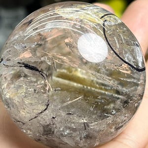 Op de afbeelding: Een heldere kwartskristallen bol met interne insluitsels en donkere lineaire markeringen. De bol heeft een gepolijst oppervlak en een reflecterende kwaliteit, met een heldere witte stip. De kristal wordt in een hand gehouden.