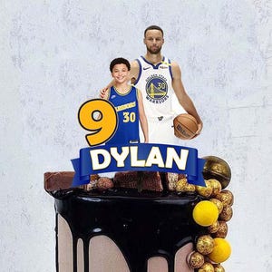 Puede incluir: Una tarta de cumpleaños con goteo de ganache de chocolate, decorada con una pancarta azul que dice "DYLAN" y un gran número 9 amarillo. La tarta presenta jugadores de baloncesto y una pelota de baloncesto. La tarta está decorada con chocolates y caramelos amarillos.