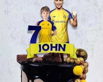 May include: A birthday cake with a chocolate ganache drip. The cake is decorated with a banner that says "JOHN", the number "7", and images of two people wearing yellow and blue soccer jerseys. The cake is also decorated with chocolate and gold candies.
