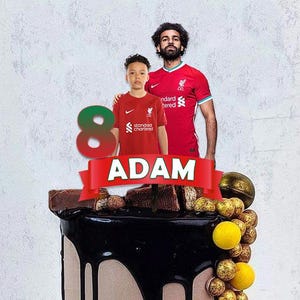 May include: A chocolate cake decorated with a red number 8, a red and white banner with the name "ADAM", and a photo of two people wearing red soccer jerseys. The cake is decorated with chocolate pieces and gold and yellow candy.
