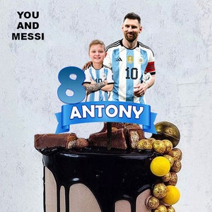 Puede incluir: Un pastel de chocolate decorado con una pancarta azul y blanca que dice "ANTONY" y un número 8 azul. El pastel está cubierto con barras de chocolate, chispas de chocolate y caramelos dorados y amarillos. Dos figuras están en el pastel, una vistiendo una camiseta de fútbol azul y blanca con el número 10 y la otra vistiendo una camiseta de fútbol azul y blanca con el texto "YOU AND MESSI".