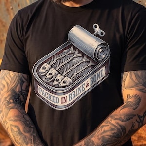 May include: A black t-shirt featuring a graphic of an open metal tin containing fish skeletons. The illustration includes a winding key on the lid and the text "PACKED IN BRINE & BONE" printed in a curved banner along the bottom of the tin design.