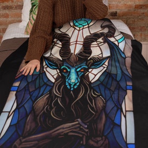 May include: A black fleece blanket with a stained glass design of a blue and black goat with horns and wings. The goat is sitting on a throne with a crown on its head.