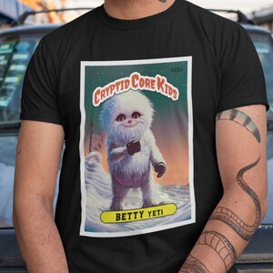Op de afbeelding: Een zwart T-shirt met een witte afbeelding van een cartoon-yeti die een beker vasthoudt. De afbeelding is in de stijl van een vintage verzamelkaart met de tekst "Cryptid Core Kids" en "Betty Yeti".