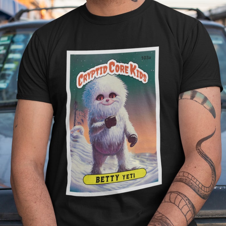 Op de afbeelding: Een zwart T-shirt met een witte afbeelding van een cartoon-yeti die een beker vasthoudt. De afbeelding is in de stijl van een vintage verzamelkaart met de tekst "Cryptid Core Kids" en "Betty Yeti".