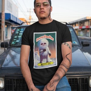 Op de afbeelding: Een zwart T-shirt met een grafische print van een cartoon Yeti. De afbeelding is een afbeelding in trading card-stijl met de tekst "Cryptid Core Kids" en "Betty Yeti".