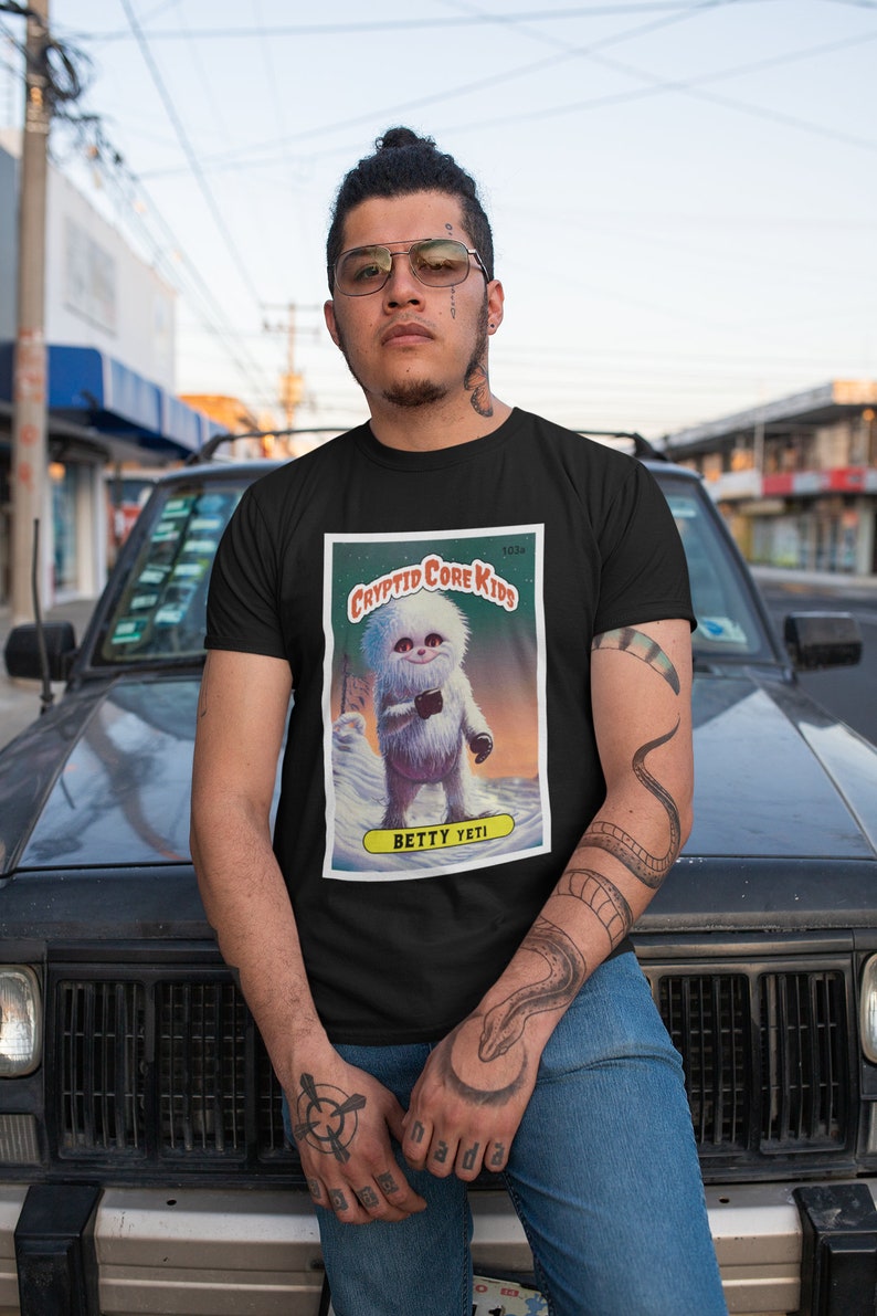 Op de afbeelding: Een zwart T-shirt met een grafische print van een cartoon Yeti. De afbeelding is een afbeelding in trading card-stijl met de tekst "Cryptid Core Kids" en "Betty Yeti".