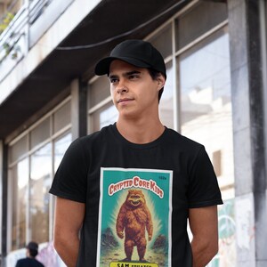 May include: Black t-shirt with a vintage-style illustration of a Bigfoot creature. The illustration is on a card with the text "Cryptid Core Kids" and "Sam Squatch".