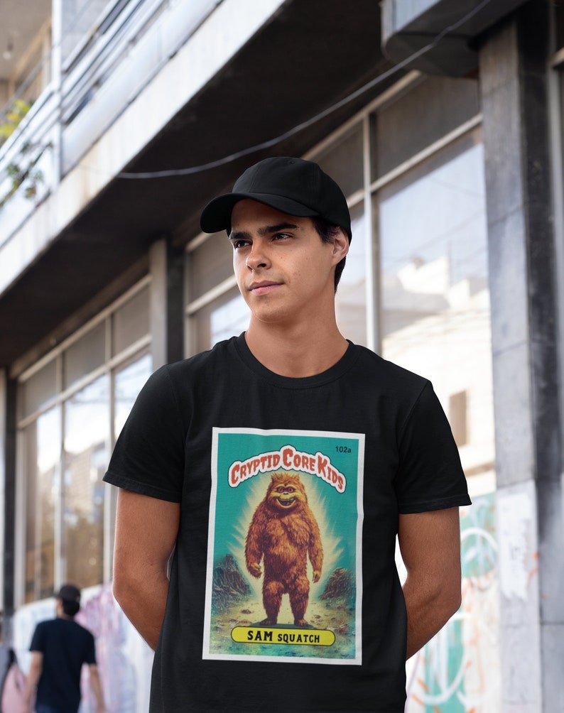 May include: Black t-shirt with a vintage-style illustration of a Bigfoot creature. The illustration is on a card with the text "Cryptid Core Kids" and "Sam Squatch".