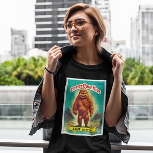 May include: A black t-shirt with a vintage-style graphic of a Bigfoot creature. The graphic is titled "Cryptid Core Kids" and features the text "Sam Squatch" at the bottom.