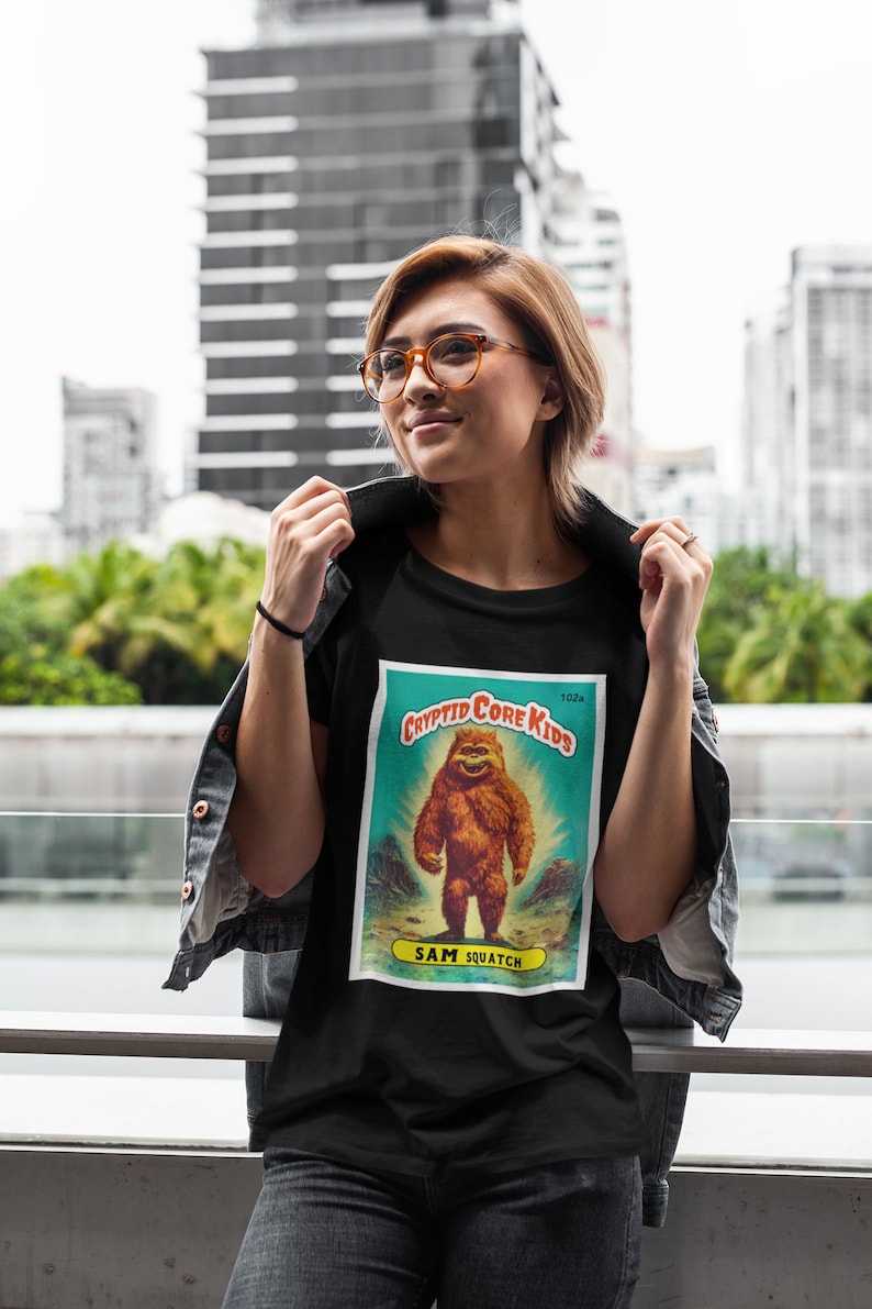 May include: A black t-shirt with a vintage-style graphic of a Bigfoot creature. The graphic is titled "Cryptid Core Kids" and features the text "Sam Squatch" at the bottom.