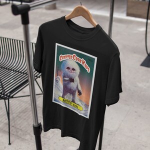 Op de afbeelding: Zwart T-shirt met een afbeelding van een Yeti-cartoon op een verzamelkaart. De kaart heeft de tekst "Cryptid Core Kids" en "Betty Yeti".