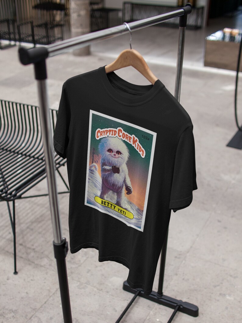 Op de afbeelding: Zwart T-shirt met een afbeelding van een Yeti-cartoon op een verzamelkaart. De kaart heeft de tekst "Cryptid Core Kids" en "Betty Yeti".