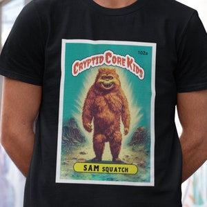 May include: A vintage-style trading card featuring a cartoon Bigfoot with a toothy grin. The card is titled "Cryptid Core Kids" and features the text "Sam Squatch" at the bottom. The card is set against a blue and green background with a light shining behind the creature.