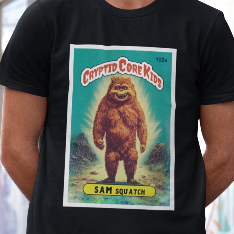 May include: A vintage-style trading card featuring a cartoon Bigfoot with a toothy grin. The card is titled "Cryptid Core Kids" and features the text "Sam Squatch" at the bottom. The card is set against a blue and green background with a light shining behind the creature.