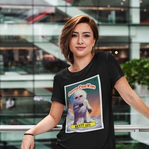 Op de afbeelding: Zwart T-shirt met een afbeelding van een witte yeti die een beker vasthoudt. De afbeelding is een vintage stijl verzamelkaart met de tekst "Cryptid Core Kids" en "Betty Yeti".