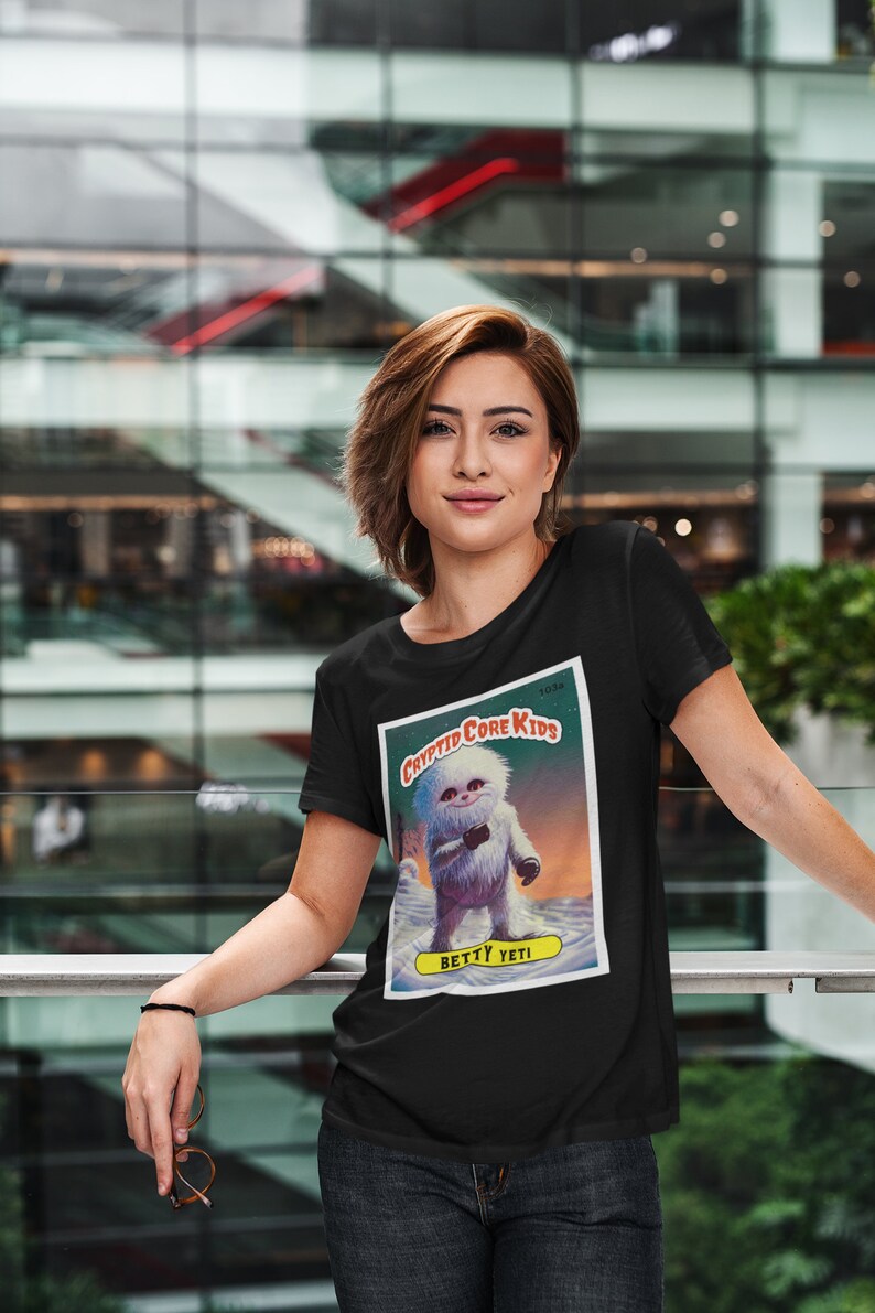 Op de afbeelding: Zwart T-shirt met een afbeelding van een witte yeti die een beker vasthoudt. De afbeelding is een vintage stijl verzamelkaart met de tekst "Cryptid Core Kids" en "Betty Yeti".