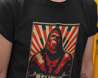Könnte beinhalten: Ein schwarzes T-Shirt mit einer roten, weißen und gelben Grafik eines Bigfoots mit dem Wort "BELIEVE" darunter.