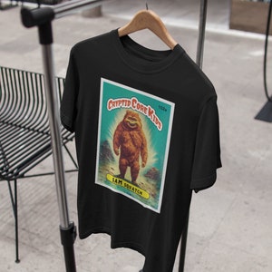 May include: Black t-shirt with a vintage-style illustration of a Bigfoot creature. The illustration is on a blue and green card with the text "Cryptid Core Kids" and "Sam Squatch".