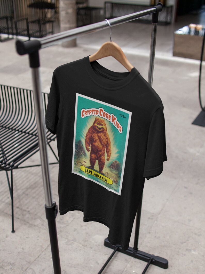 May include: Black t-shirt with a vintage-style illustration of a Bigfoot creature. The illustration is on a blue and green card with the text "Cryptid Core Kids" and "Sam Squatch".