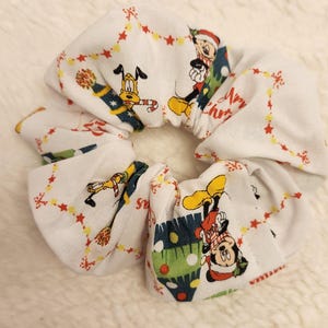 May include: White scrunchie with a Christmas-themed print featuring Mickey Mouse and Pluto. The print includes red and green Christmas trees, stars, and the words "Merry Christmas".