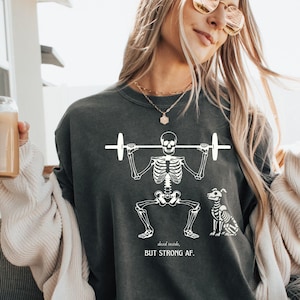 May include: A woman wearing a dark grey sweatshirt with a white skeleton lifting weights and a dog beside it. The text on the shirt reads "dead inside, but strong af."