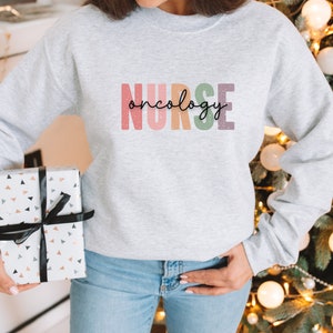 K&ouml;nnte beinhalten: Ein grauer Pullover mit dem Text "NURSE oncology" in Regenbogenfarben. Der Text ist in einer stilisierten Schriftart mit schwarzem Umriss.