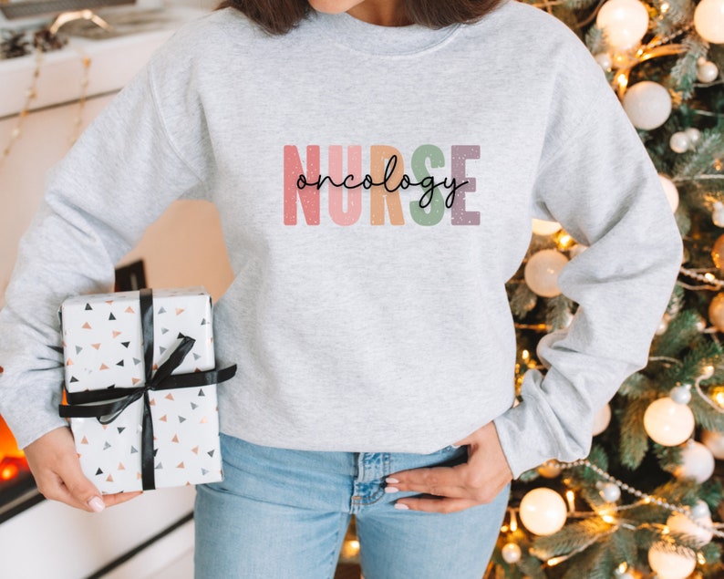 K&ouml;nnte beinhalten: Ein grauer Pullover mit dem Text "NURSE oncology" in Regenbogenfarben. Der Text ist in einer stilisierten Schriftart mit schwarzem Umriss.