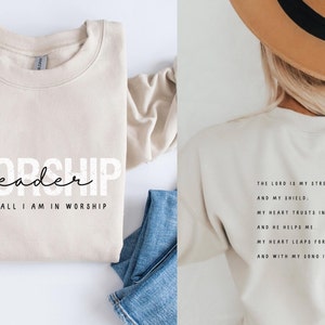 Puede incluir: Sudadera beige con el texto "Worship leader" y "Giving all I am in worship" en la parte delantera. La parte posterior de la sudadera tiene el texto "The Lord is my strength and my shield. My heart trusts in Him, and He helps me. My heart leaps for joy, and with my song I praise Him. Psalm 28:7"