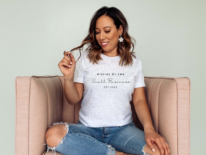 May include: A woman wearing a grey t-shirt with the text "Minding My Own Small Business Est. 2023" printed on it. She is sitting on a pink couch and smiling.