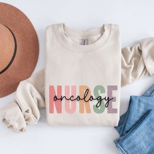 K&ouml;nnte beinhalten: Ein beiger Pullover mit dem Text "NURSE oncology" in Regenbogenfarben. Der Text ist im Vintage-Stil mit einem gesprenkelten Hintergrund.