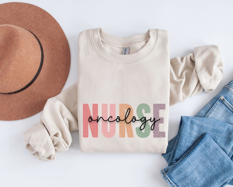 K&ouml;nnte beinhalten: Ein beiger Pullover mit dem Text "NURSE oncology" in Regenbogenfarben. Der Text ist im Vintage-Stil mit einem gesprenkelten Hintergrund.
