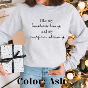 May include: A grey sweatshirt with the text "I like my lashes long and my coffee strong" printed on the front. The sweatshirt is being modelled by a person wearing blue jeans. The colour of the sweatshirt is ash.