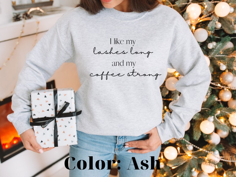 May include: A grey sweatshirt with the text "I like my lashes long and my coffee strong" printed on the front. The sweatshirt is being modelled by a person wearing blue jeans. The colour of the sweatshirt is ash.