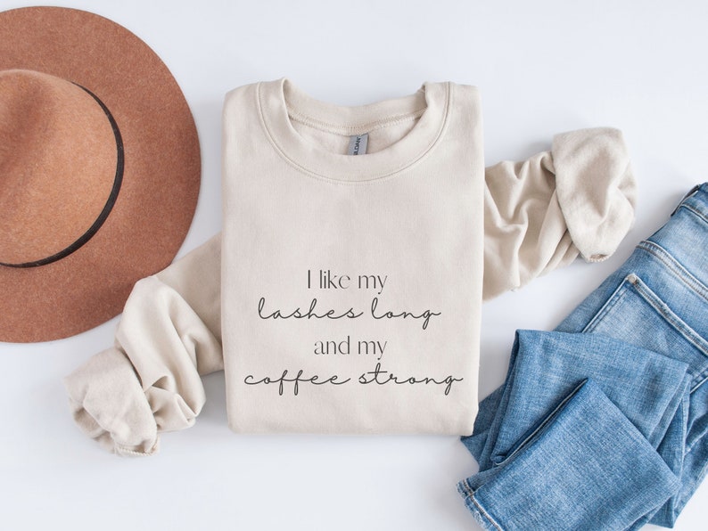 May include: A beige sweatshirt with the text "I like my lashes long and my coffee strong" printed on the front. The sweatshirt is folded and laid out on a white surface with a brown hat and blue jeans.
