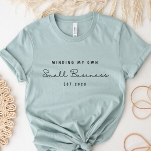 May include: A light blue t-shirt with the text "Minding My Own Small Business Est. 2023" printed in black.