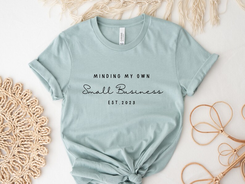 May include: A light blue t-shirt with the text "Minding My Own Small Business Est. 2023" printed in black.